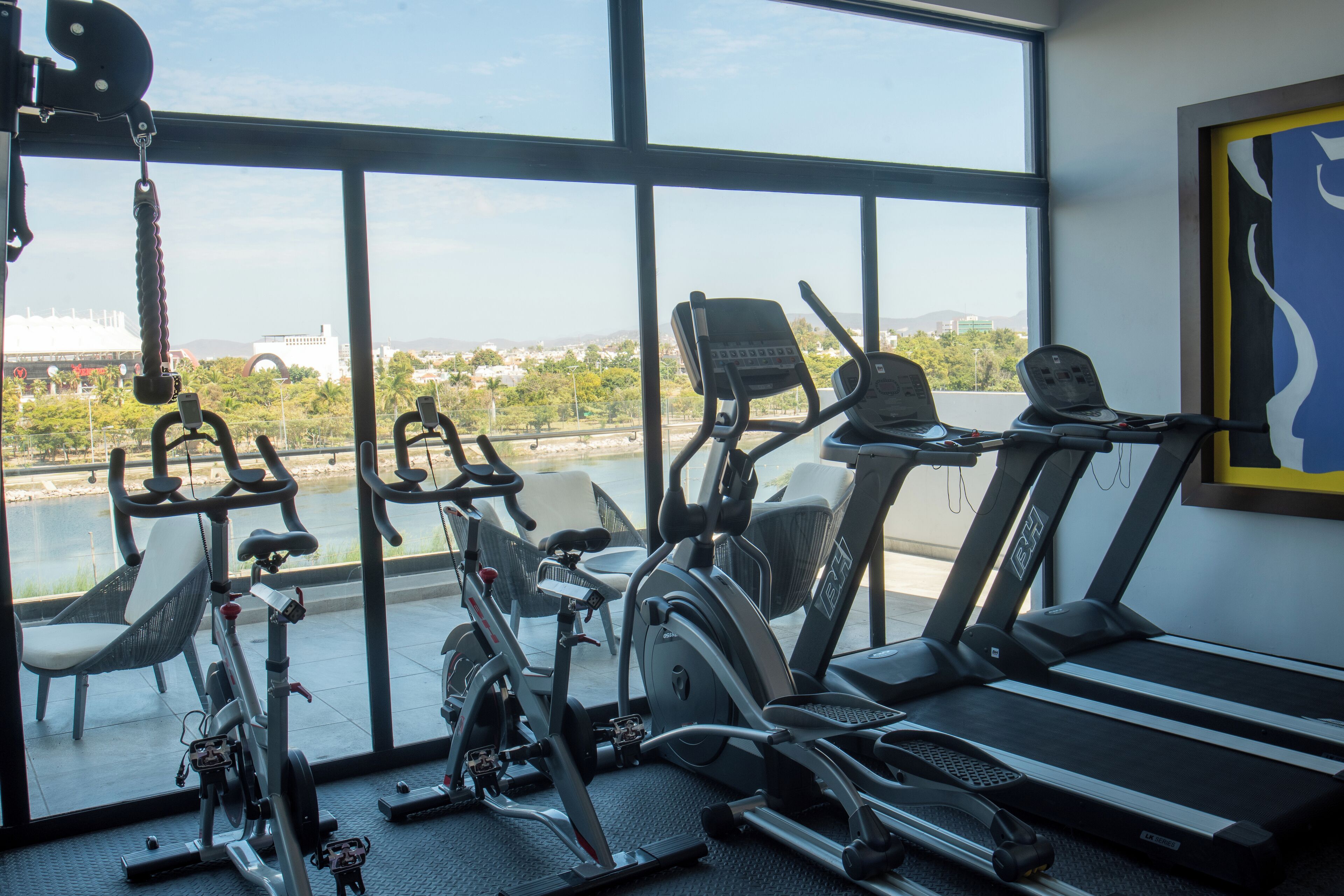 A gym with various exercise equipment, including stationary bikes and treadmills, with a view of a cityscape through large windows.