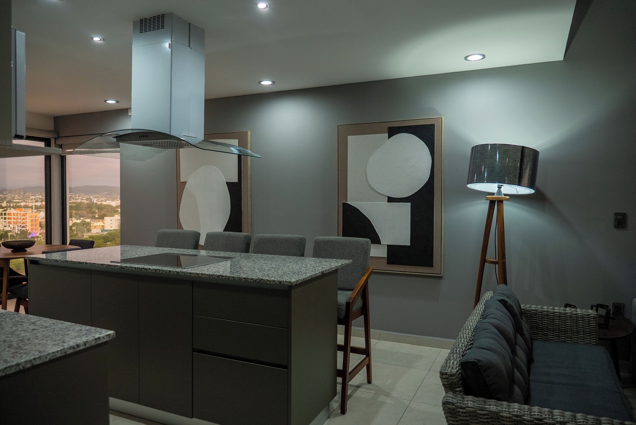 A modern kitchen with a central island, a dining area with chairs, a standing lamp, and abstract wall art.