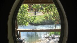 A view through a circular window of a riverside area with a wooden deck and a small plant in the foreground. - Sleeps 25, 5 Homes, Fire Pit, Spring Fed Waterfront (Ingram)