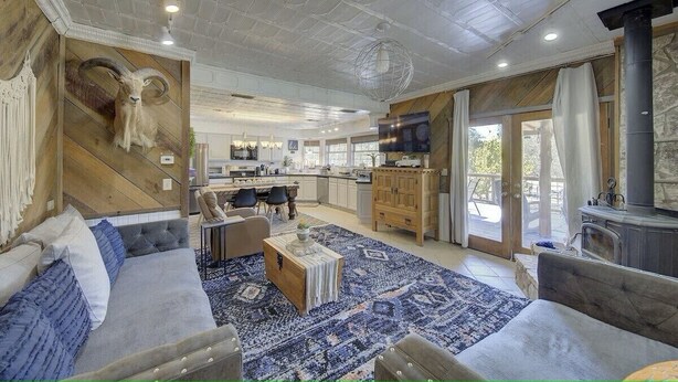 A spacious living room with a large sofa, a coffee table, and a mounted ram skull. There is a kitchen area with a stove and a dining table. The room has large windows and a stone fireplace. - Sleeps 25, 5 Homes, Fire Pit, Spring Fed Waterfront (Ingram)