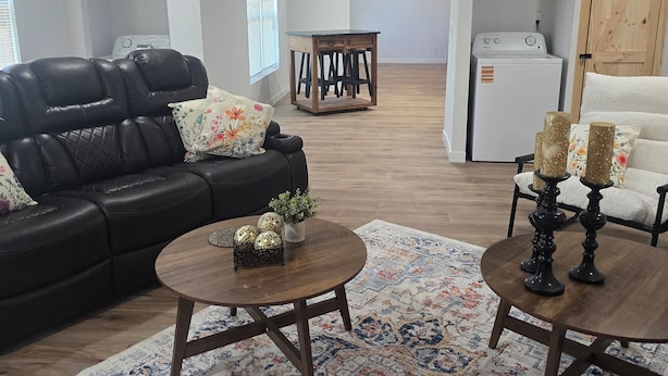 A living room with a black leather sofa, a wooden coffee table, a patterned rug, a ceiling fan, and a laundry area with a washing machine.