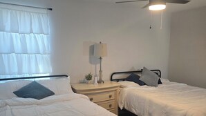 A bedroom with two beds, a nightstand, a lamp, a ceiling fan, and a window with curtains.
