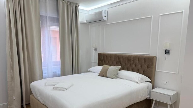 A neatly made bed with white linens, a wooden headboard, and a small bedside table with a lamp. - Emy Suites (Civitavecchia)