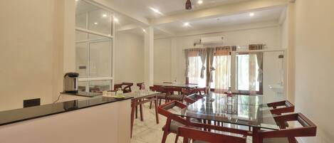 A spacious dining area with a glass-top table and wooden chairs, a kitchen counter, and a large window with curtains.