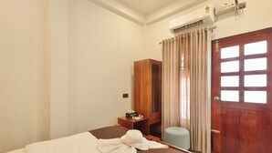 Une chambre d’hôtel comprenant un lit, une armoire en bois, un bureau avec un miroir, un petit tabouret et une porte avec une fenêtre.