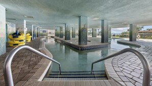 An indoor swimming pool with clear water, surrounded by tiled walkways and supported by pillars. - New Direct Ocean View 12th Floor, Sandcastle South (Myrtle Beach)