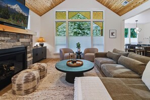 Ein geräumiges Wohnzimmer mit Kamin aus Stein, einem runden Couchtisch und einem großen Fenster mit Blick auf Grünflächen.