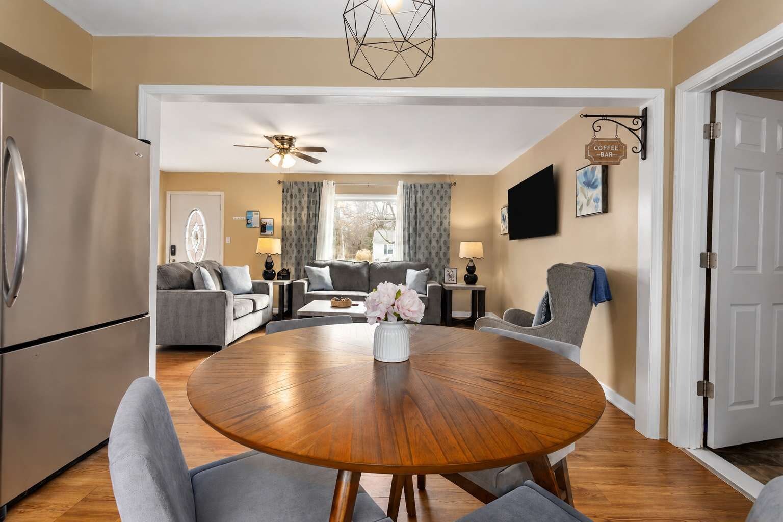 A modern living room with a round wooden dining table, grey sofas, a ceiling fan, and a kitchen area with a refrigerator.