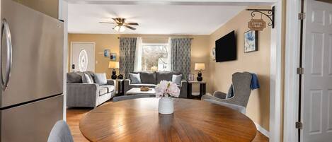 A modern living room with a round wooden dining table, grey sofas, a ceiling fan, and a kitchen area with a refrigerator.