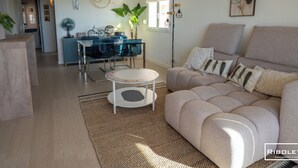 Ett modernt vardagsrum med en beige soffa, ett matbord i glas och ett köksområde i bakgrunden.