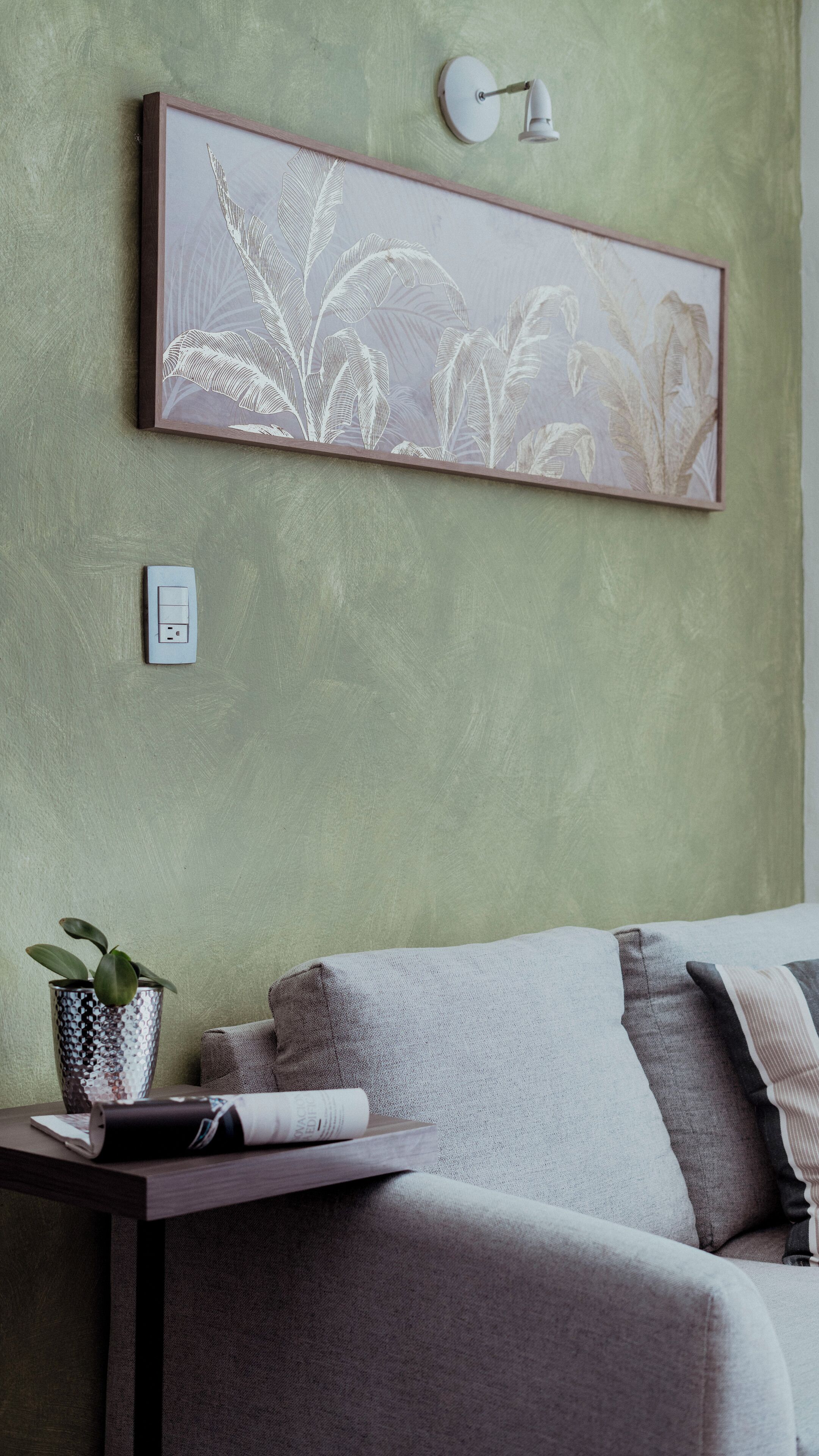 A room with a green textured wall, a framed botanical print, a bedside table with a plant, and a lamp.