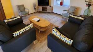 Ein Wohnzimmer mit Holzboden, zwei schwarzen Sofas mit gemusterten Kissen, einem Holztisch, einem Fernseher auf einem Schrank und einem Fenster mit Blick.