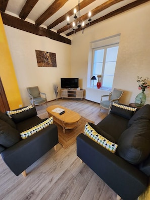 A living room with a wooden floor, two black sofas with patterned cushions, a wooden coffee table, a TV on a stand, and a window with a view. - Maison de village entre le futuroscope et Beauval (Preuilly-sur-Claise)
