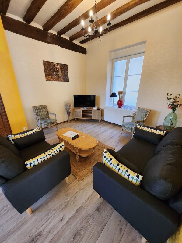 Un soggiorno con pavimento in legno, due divani neri con cuscini decorati, un tavolino in legno, una TV su un mobile basso e una finestra con vista.