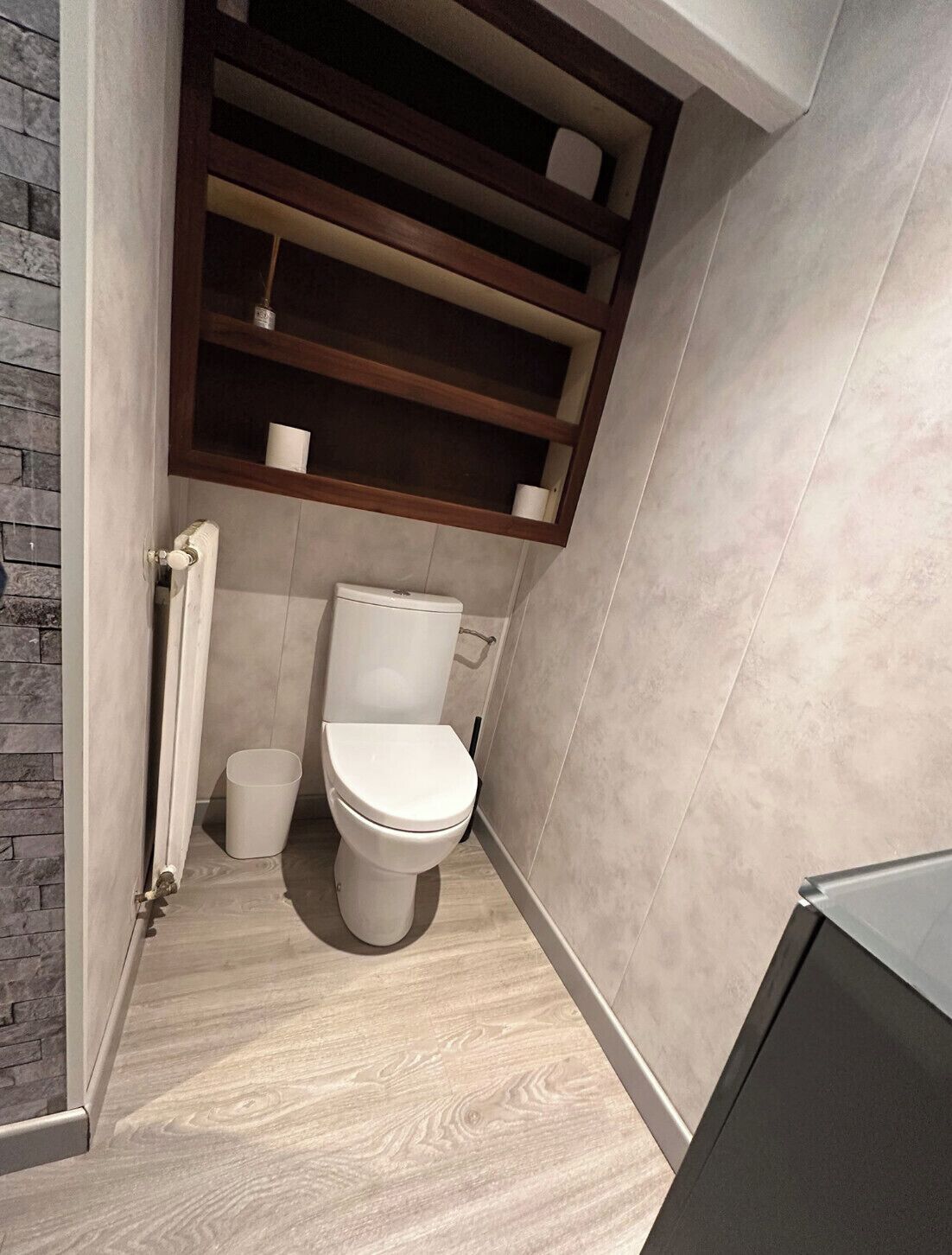 Une petite salle de bain comprenant des toilettes, une petite poubelle et une étagère avec une tasse et un rouleau de papier toilette.