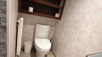 Une petite salle de bain comprenant des toilettes, une petite poubelle et une étagère avec une tasse et un rouleau de papier toilette.