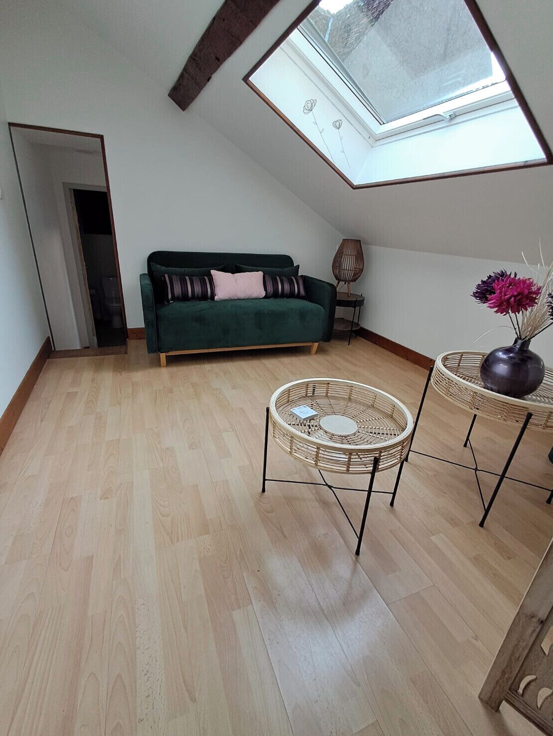 Et loft-studie med ovenlys, en grøn sofa, to fletvævetænger og en vase med blomster.