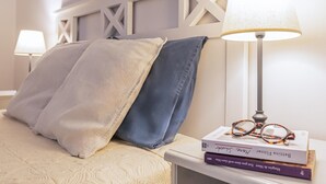 Ein ordentlich bezogenes Bett mit beigem und blauem Kopfkissen, ein weißer, geometrisch gestalteter Kopfteil und ein Nachttisch mit Lampe und Büchern.