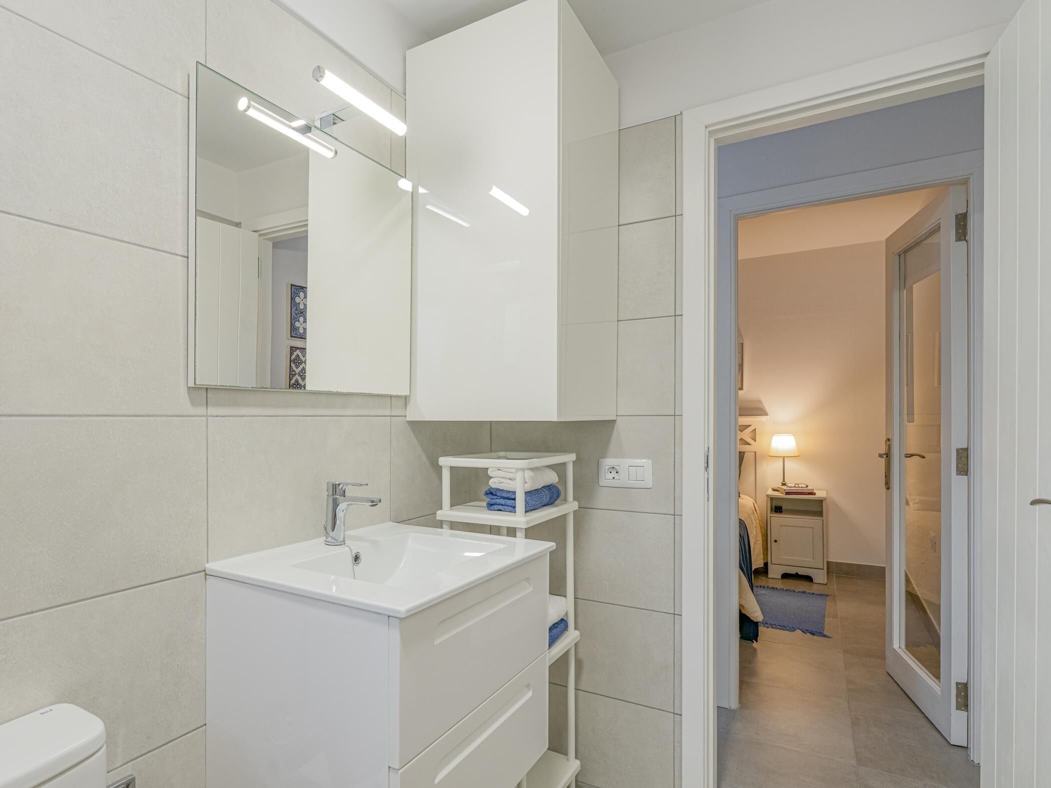 Une salle de bain moderne avec un lavabo blanc, un miroir et un meuble, et une porte visible donnant sur une autre pièce.