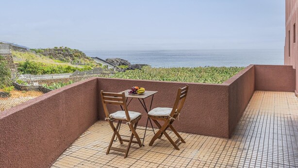 Ein Balkon mit zwei Holzstühlen und einem kleinen Tisch, der Blick auf die Küste mit blauem Himmel bietet.