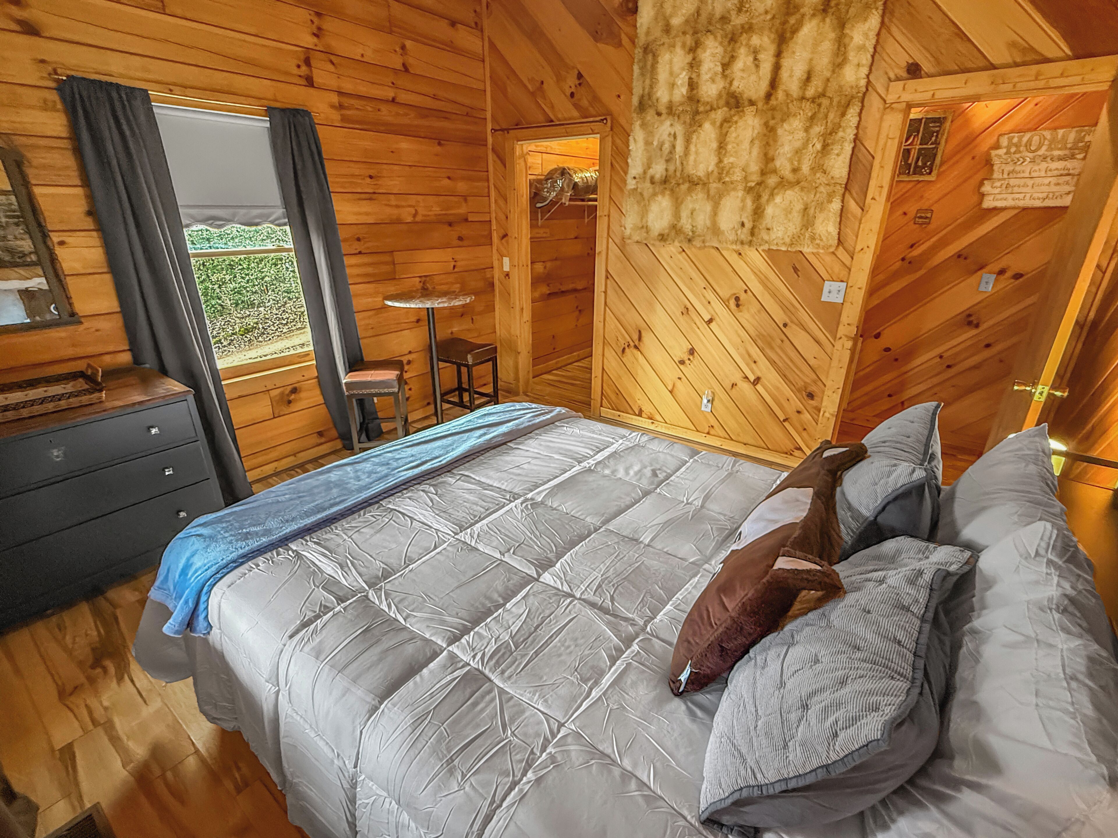 A wooden cabin bedroom with a bed, a dresser, a small table, and a window with curtains.