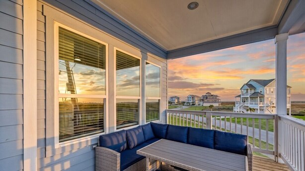 Ein überdachter Balkon mit Sitzgelegenheit, großen Fenstern und Blick auf das Meer sowie umliegende Häuser.