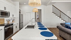 Eine moderne Küche mit weißen Schränken, einer zentralen Kochinsel mit blauen Platzsets, einer großen gewebten Pendelleuchte und einer Treppe, die zu einer anderen Ebene führt.