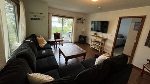 Ein Wohnzimmer mit einem schwarzen Ecksofa, einem Holztisch, einem Fernseher und einer Glastür, die auf eine Außenpatio führt.