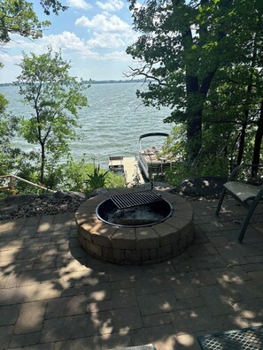 Un hogar circular con parrilla, rodeado de una zona pavimentada con vistas a un lago y un muelle.