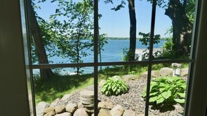 Blick durch ein Fenster auf eine Steinmauer, einen kleinen Garten mit Pflanzen und ein Gewässer im Hintergrund.