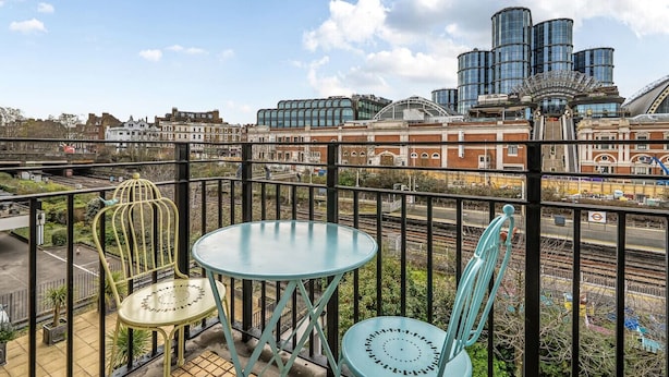 Un balcon avec des chaises et une table en métal, donnant sur une voie ferrée et des bâtiments modernes.