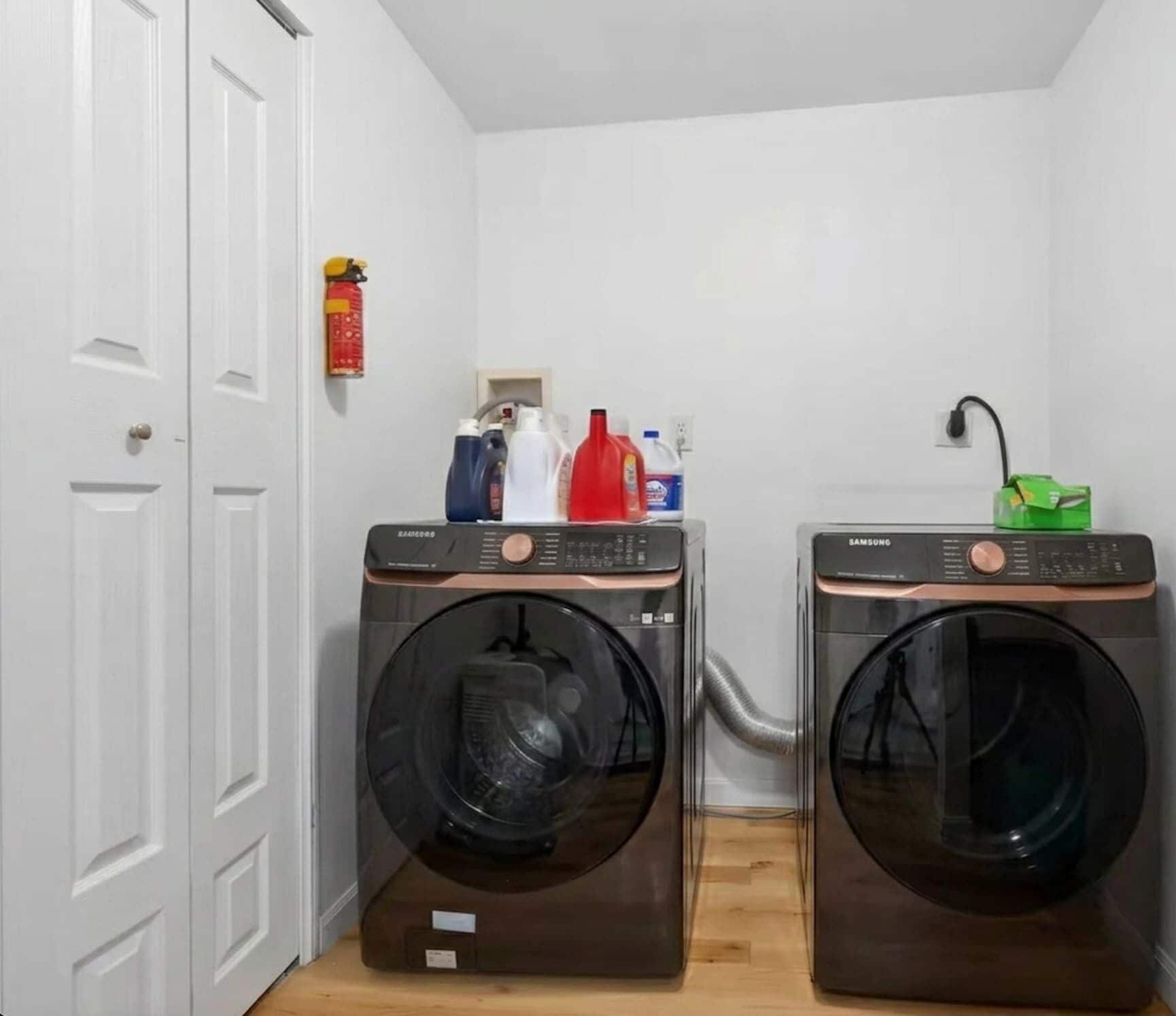 A laundry room with two front-loading washing machines, a fire extinguisher, and cleaning supplies on a shelf.
