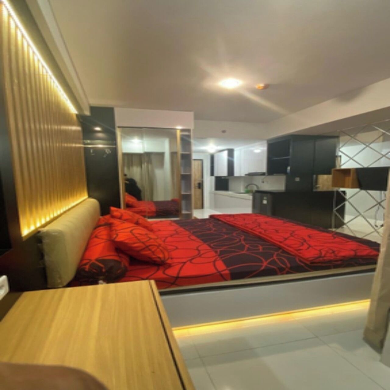 A modern hotel room with a large bed, a wooden desk, a built-in kitchen, and a balcony.