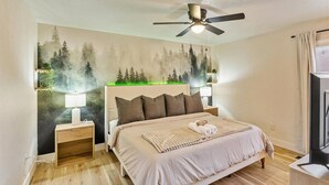 A bedroom with a large bed, a ceiling fan, a nightstand with a lamp, and a wall mural of a forest. - Hot Tub Getaway and Quiet Home Near Downtown SLC (South Salt Lake)