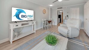 A modern living room with a television displaying 'BIG FISH Rentals,' a white console table, a white sofa, a dining area with chairs, and a ceiling fan. - Decorated 1BR Condo at Ocean Dunes Tower 1 with Pool (Myrtle Beach)