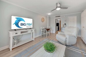 A modern living room with a television displaying 'BIG FISH Rentals,' a white console table, a white sofa, a dining area with chairs, and a ceiling fan. - Decorated 1BR Condo at Ocean Dunes Tower 1 with Pool (Myrtle Beach)