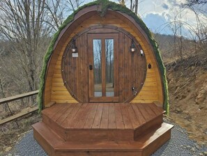 Una cabaña de madera con un amplio ventanal central, rodeada por una terraza de madera, ubicada en un paisaje natural.