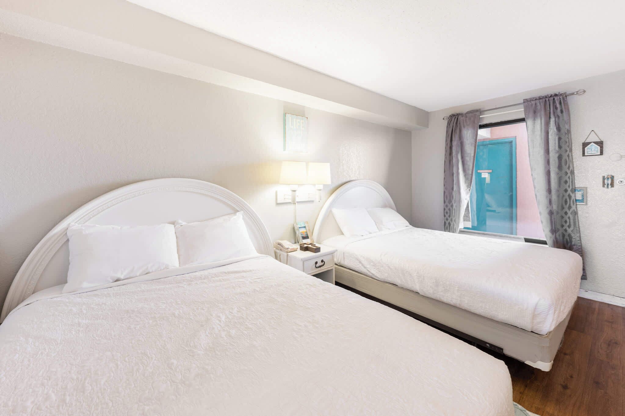 Two beds with white bedding, a bedside table with a lamp, and a window with curtains.