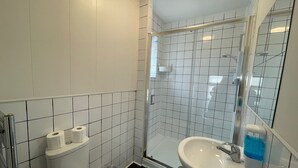 Uma casa de banho pequena com azulejos brancos, sanita, lavatório e chuveiro.