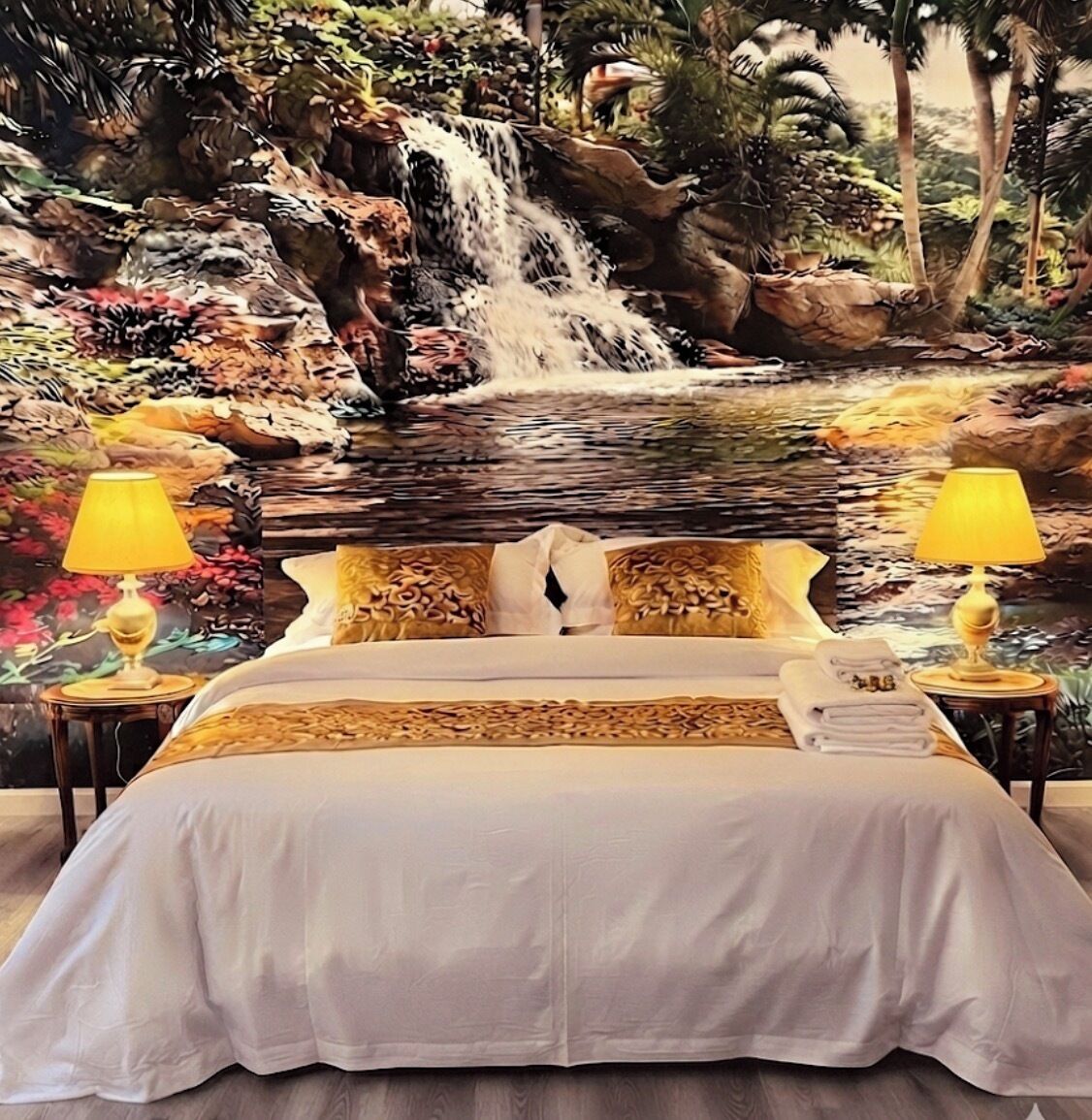 A bed with a white bedspread, two bedside lamps, and a view of a waterfall and foliage.