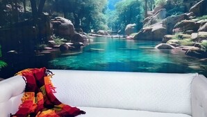 Ein weißes Sofa mit einer bunten Decke, ein klares, blaues Schwimmbecken und eine natürliche, bewaldete Gegend.