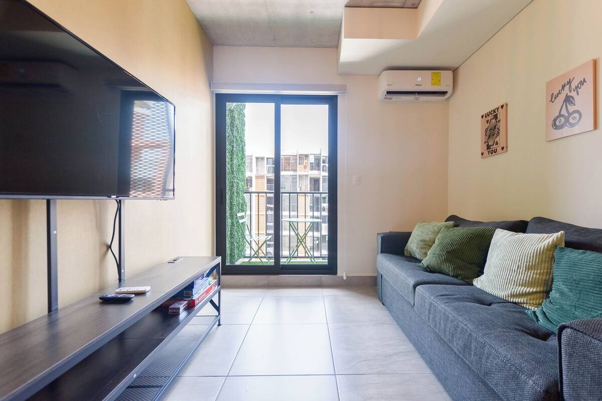 A living room with a flat-screen TV, a sofa with cushions, a small table, and a sliding glass door leading to a balcony.