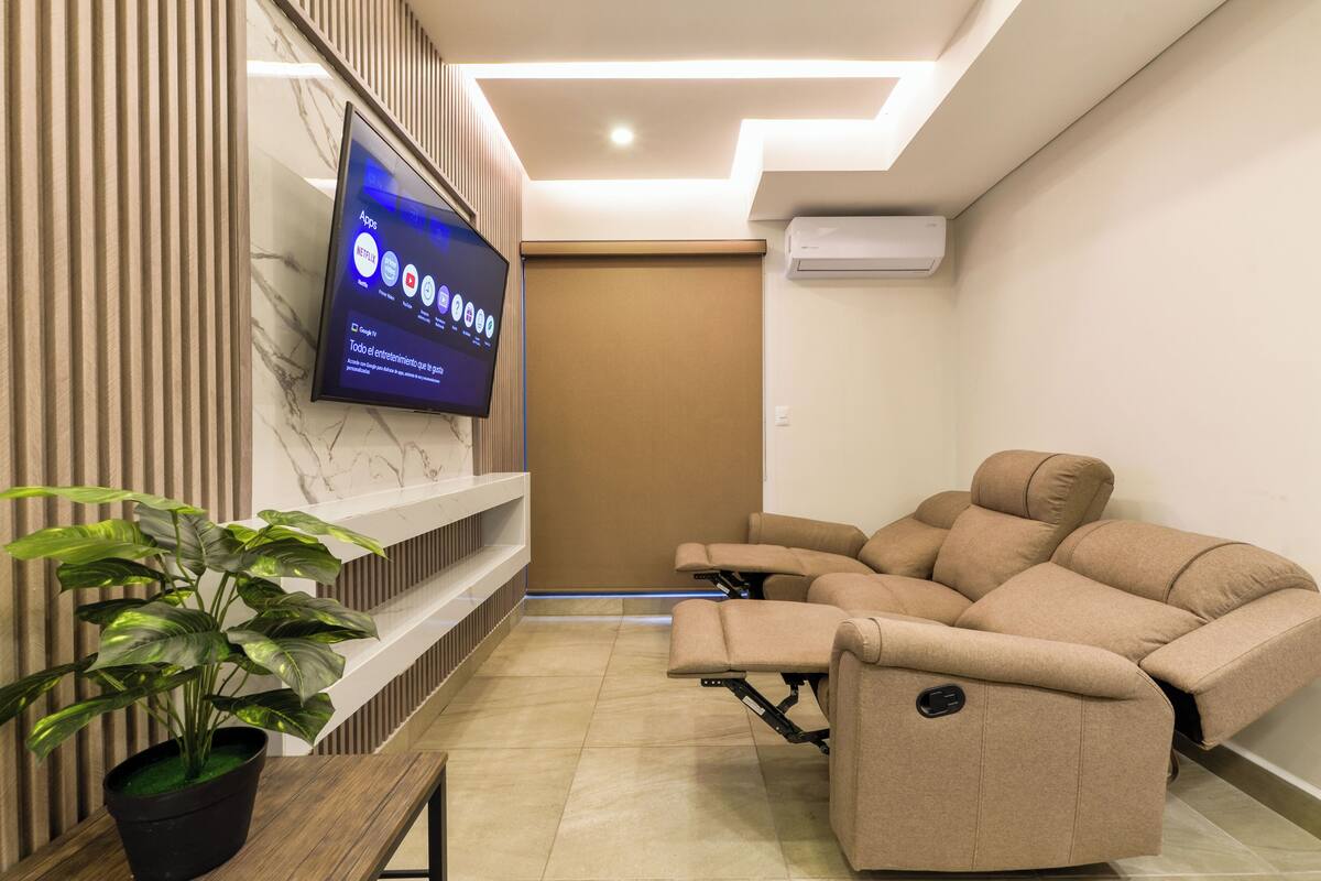 A modern living room with a flat-screen TV mounted on a wall, a reclining sofa, a potted plant, and a built-in shelf.