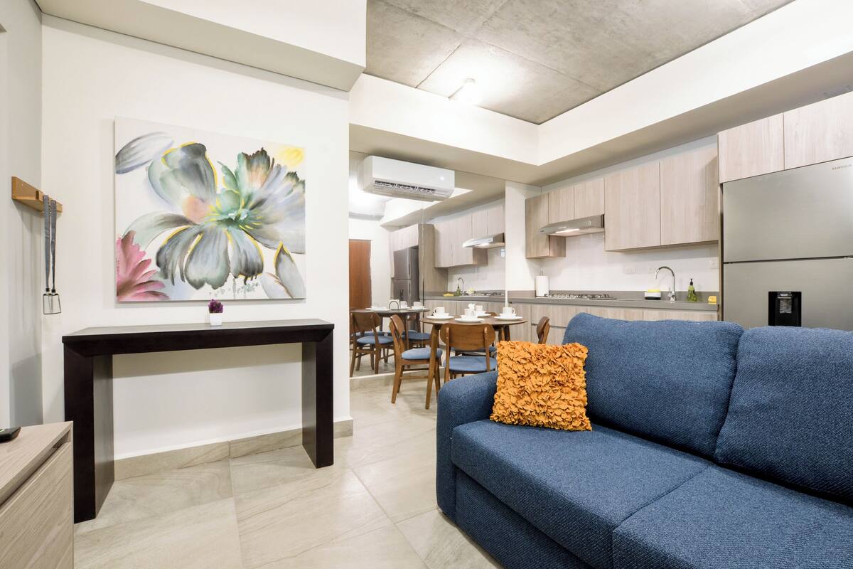 A modern living room with a blue sofa, a dining area with wooden chairs, and a kitchen with light wooden cabinets and stainless steel appliances.