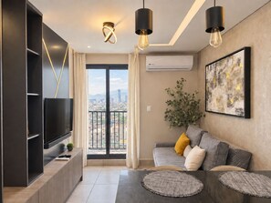 A modern living room with a flat-screen TV, a sofa with cushions, a large window with a city view, and a mounted air conditioner. - Monteleva (Monterrey)