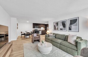 Een moderne woonkamer met een groene bank, een ronde salontafel en een eethoek met een tafel en stoelen. Achtergrond toont een keuken met donkere kastjes en een magnetron.