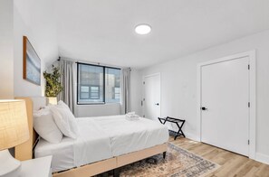 Een moderne slaapkamer met een bed, een nachtkastje, een lamp, een raam met zonwering, een deur en een kleine plant.