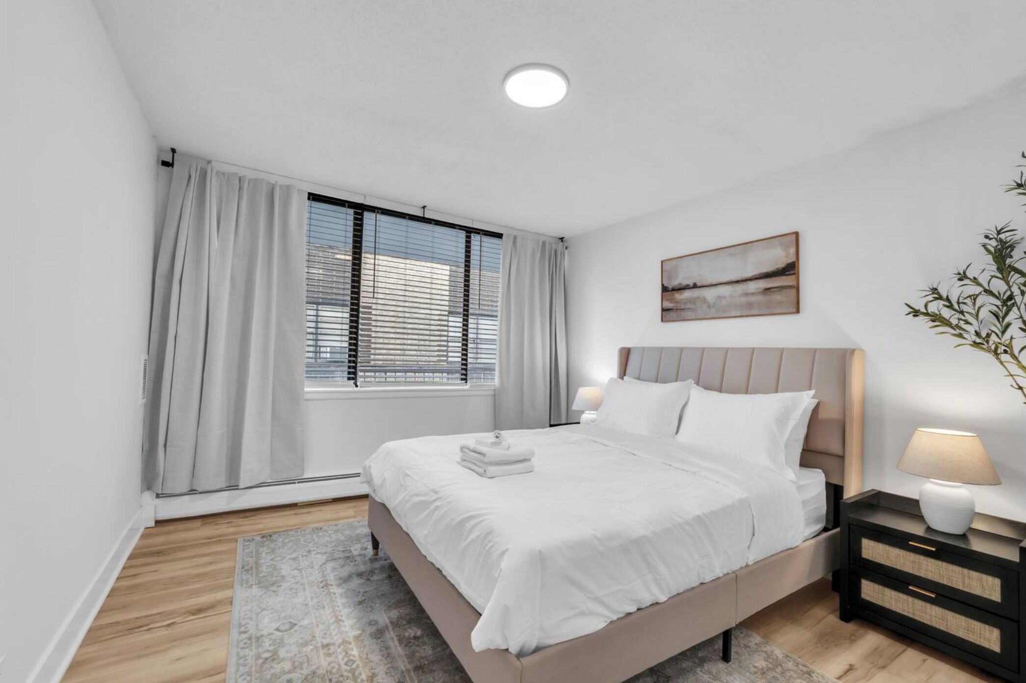 A modern bedroom with a bed, bedside tables, a window with blinds, and a painting on the wall.