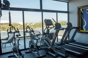 Ein Fitnessstudio mit unterschiedlichen Trainingsgeräten, darunter Fahrrädern und Laufbändern, mit Blick auf eine Stadtkulisse durch große Fenster.