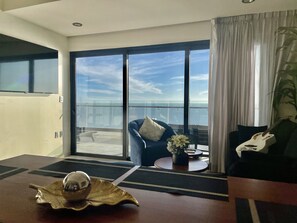 Ein Hotelzimmer mit einer großen Schiebeglasdoor zum Balkon mit Meerblick. Ein blauer Sessel mit weißem Kissen, ein kleiner runder Tisch mit Blumenvase und eine dekorative Muschel stehen auf dem Tisch.